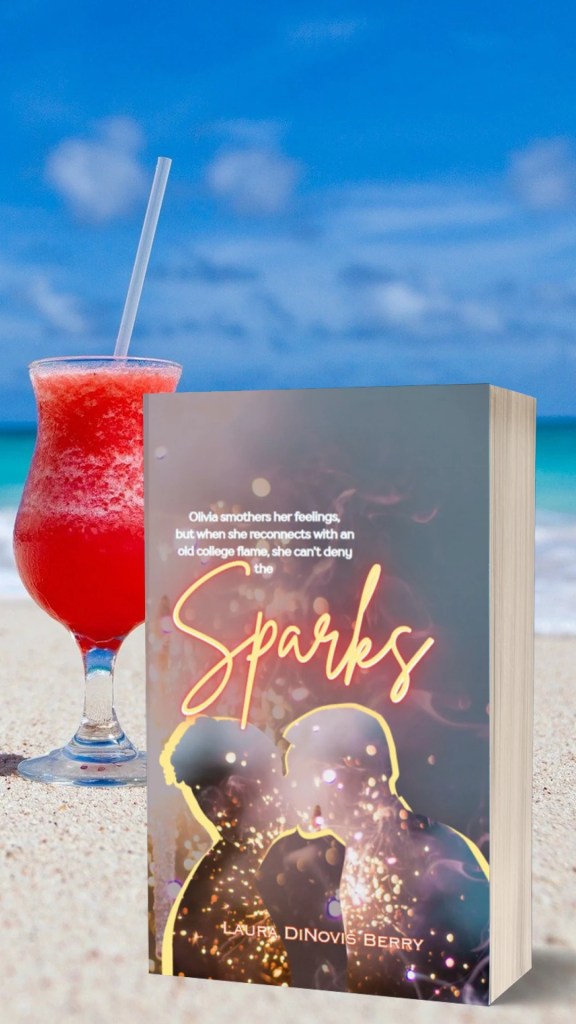 A book titled 'Sparks' by Laura Dinovis-Berry, featuring silhouettes of two people, placed on a sandy beach next to a pink tropical drink with a straw and a picturesque ocean background.
